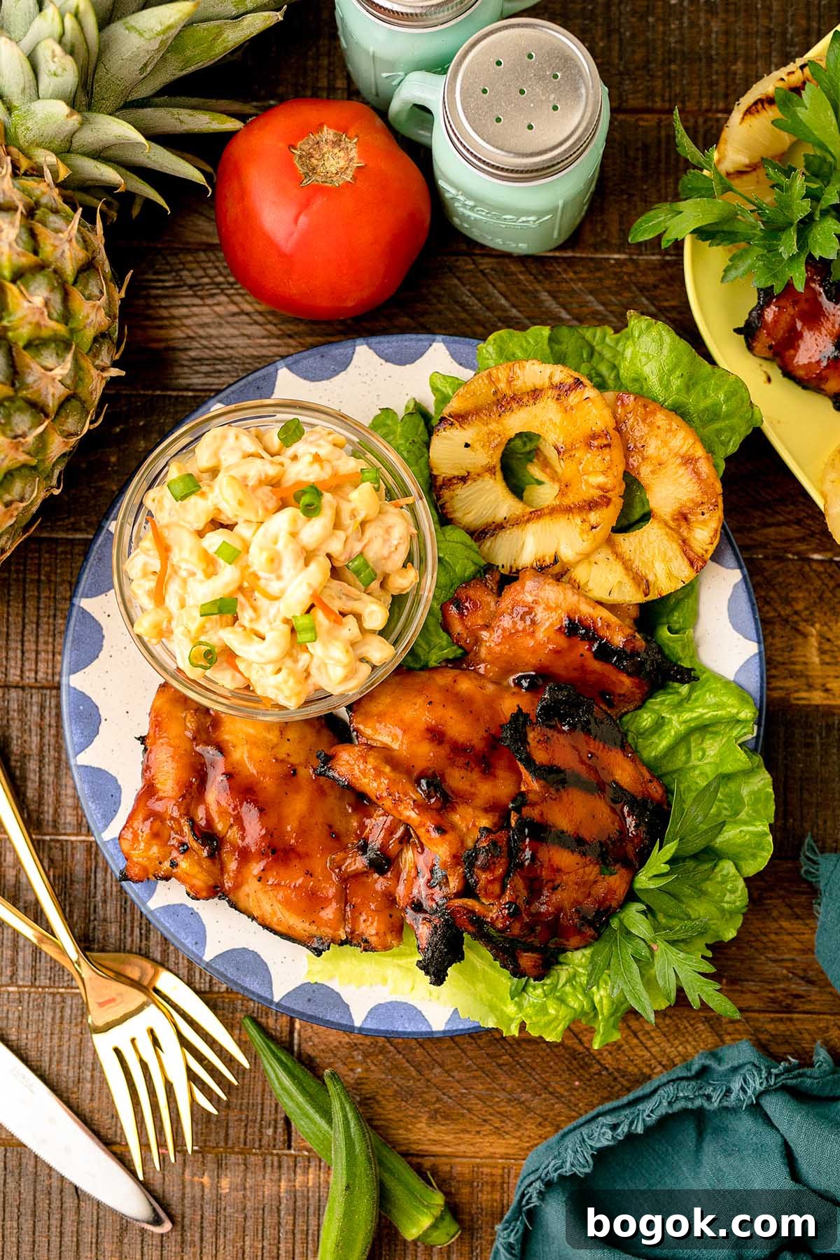 Hawaiian-Style BBQ Chicken with Pineapple Glaze 7 Savor the taste of Hawaiian BBQ Chicken with grilled chicken in barbecue sauce, accompanied by juicy sliced pineapple and a creamy macaroni salad. The vibrant plate is garnished with fresh lettuce, while the backdrop features pineapple and tomatoes for a tropical touch.