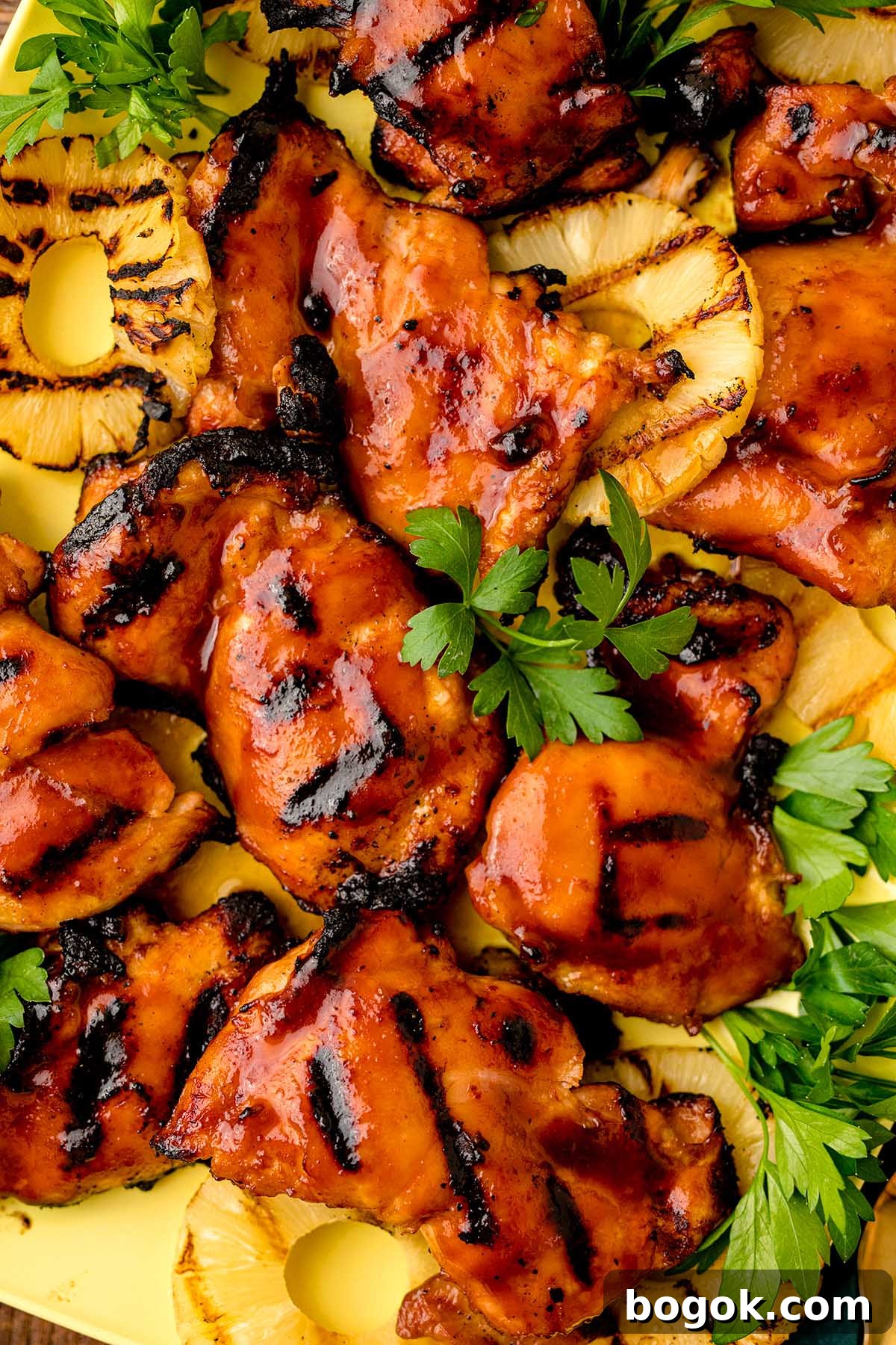Hawaiian-Style BBQ Chicken with Pineapple Glaze 9 Hawaiian BBQ chicken thighs with char marks are garnished with fresh parsley and served alongside grilled pineapple slices on a yellow plate.