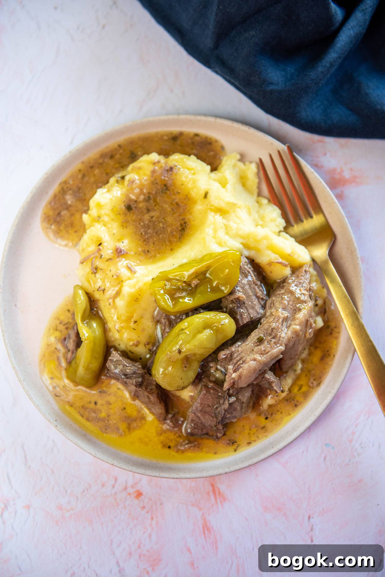 Mississippi Pot Roast Recipe for Tender Slow-Cooker Comfort 11 roast in brown liquid mixture and peperocini on top with a side of mashed potatoes served on plate on pink table with a fork on plate