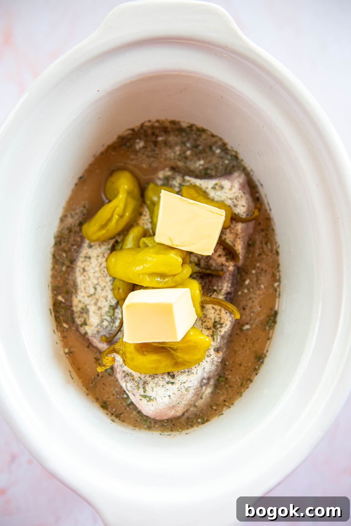 Mississippi Pot Roast Recipe for Tender Slow-Cooker Comfort 9 uncooked roast with flour and variety of spices on top, sticks of butter, peperocini in broth inside white slow cooker bowl