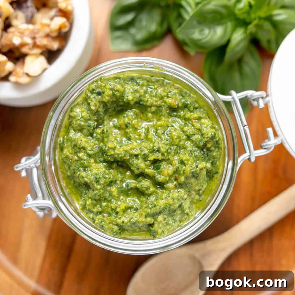 A glass jar filled with green pesto sits on a wooden surface, surrounded by fresh basil leaves, walnuts, and a wooden spoon.