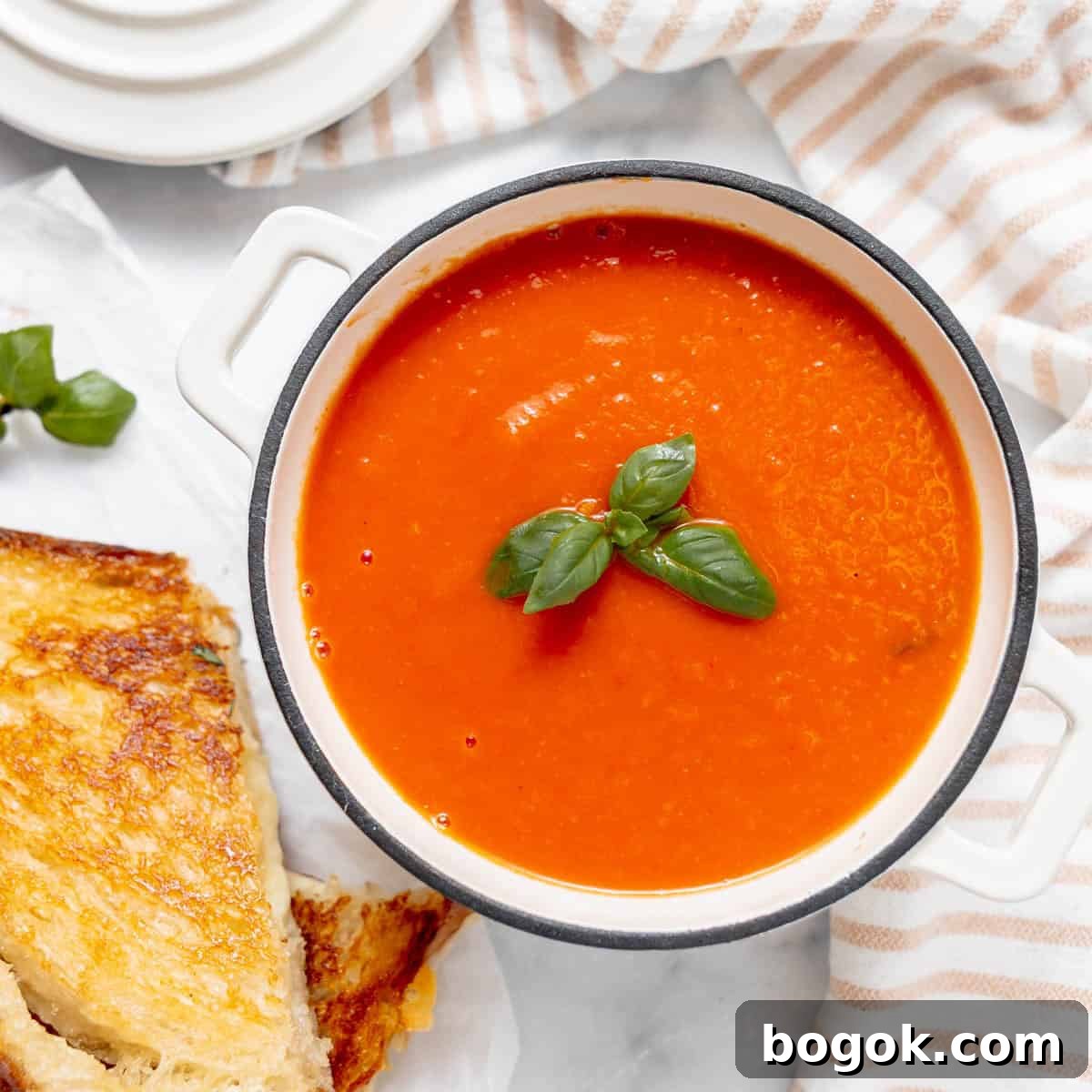 A white bowl of tomato soup garnished with fresh basil leaves, next to a grilled cheese sandwich on a white surface with a striped cloth.