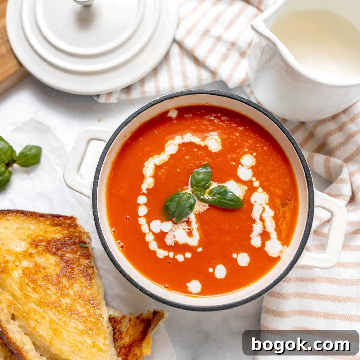 A bowl of tomato soup with cream and basil garnish, served alongside a grilled cheese sandwich on a white surface with a striped cloth napkin.