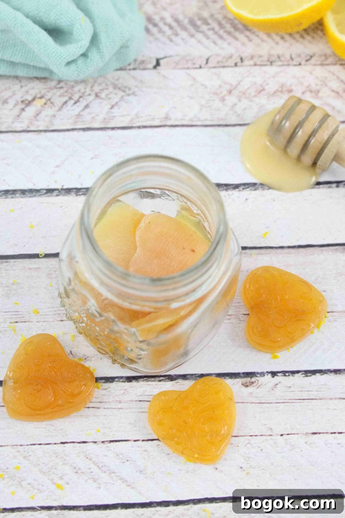 three honey lemon cough drops around a jar of more cough drops with a honey stick, fresh lemon and a blue linen behind them.
