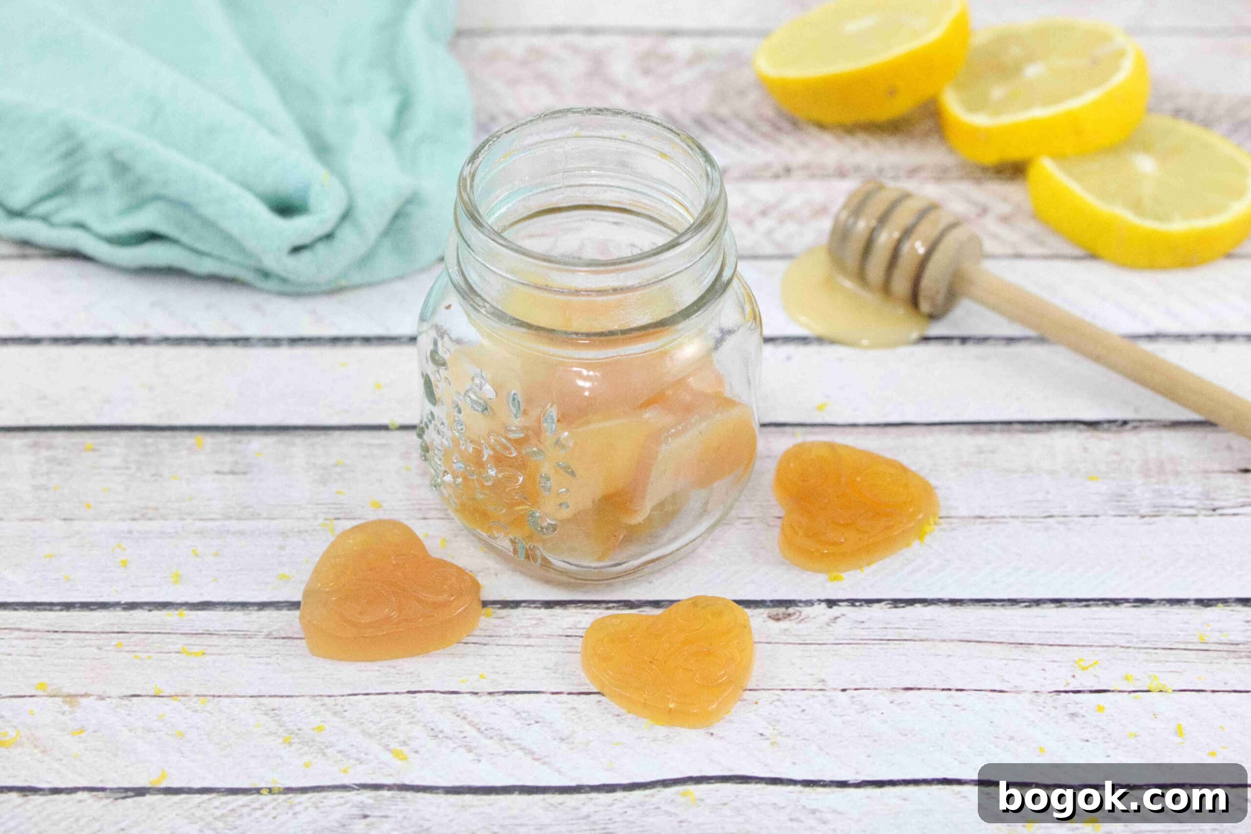 three honey lemon cough drops around a jar of more cough drops with a honey stick, fresh lemon and a blue linen behind them.