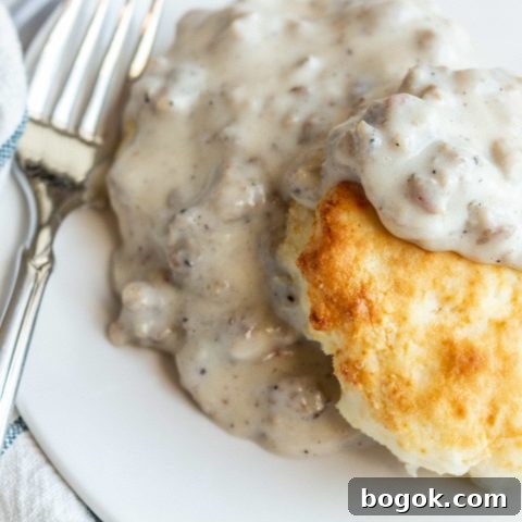 A biscuit sits on a round white plate topped with a gray sausage gravy. A fork sits angled on the side of a plate and a soft white and blue striped towel is underneath