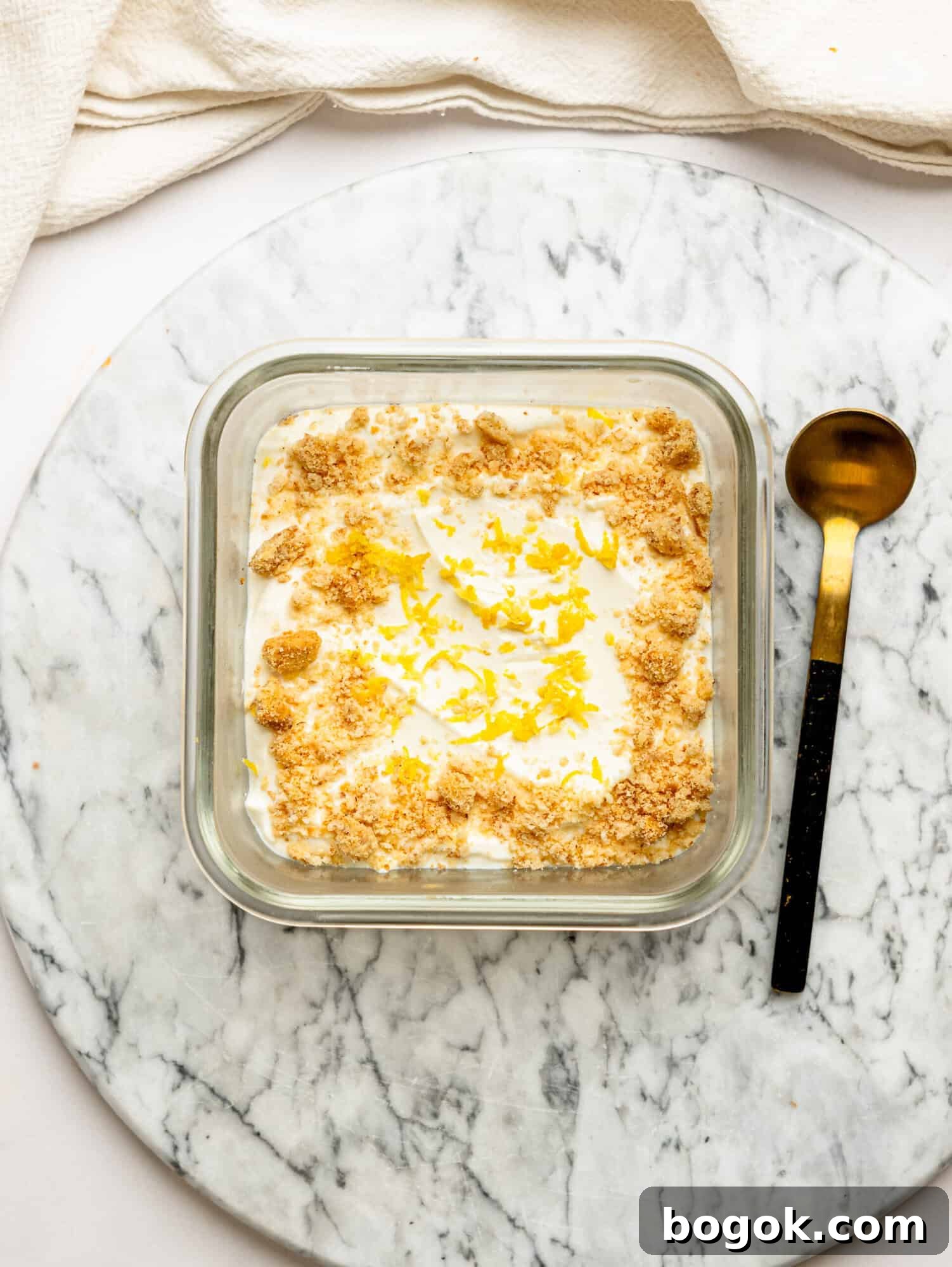 Lemon Cheesecake Overnight Weet-Bix Recipe 2 Lemon cheesecake overnight Weet-Bix in a glass container topped with biscuit crumbs and lemon zest, served on a marble board with a spoon alongside.