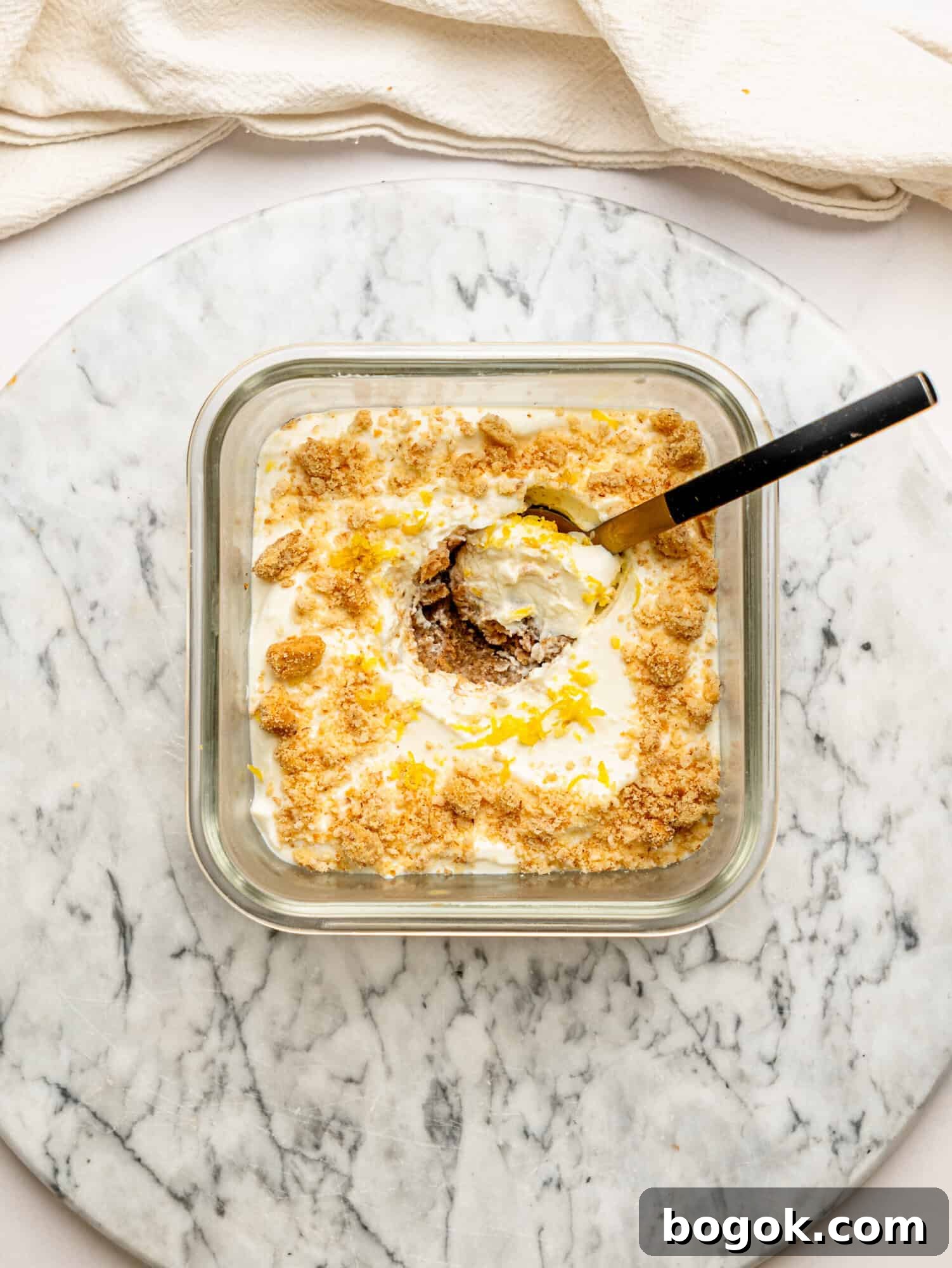 Lemon Cheesecake Overnight Weet-Bix Recipe 3 Lemon cheesecake overnight Weet-Bix in a glass meal prep container topped with crushed biscuit and fresh lemon zest, with a spoon scooping out a creamy serving.