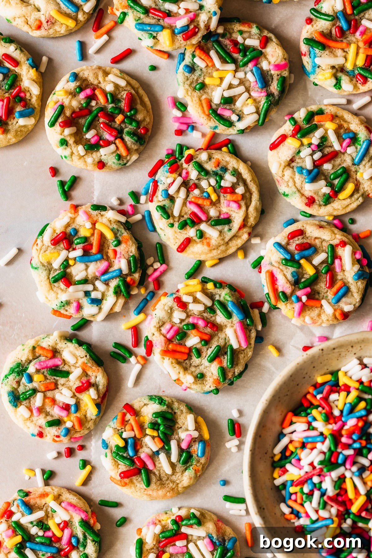 Soft and Chewy Mini Sprinkle Cookies Recipe 2 Rows of sprinkle cookies next to a white bowl of rainbow sprinkles on a pink counter.