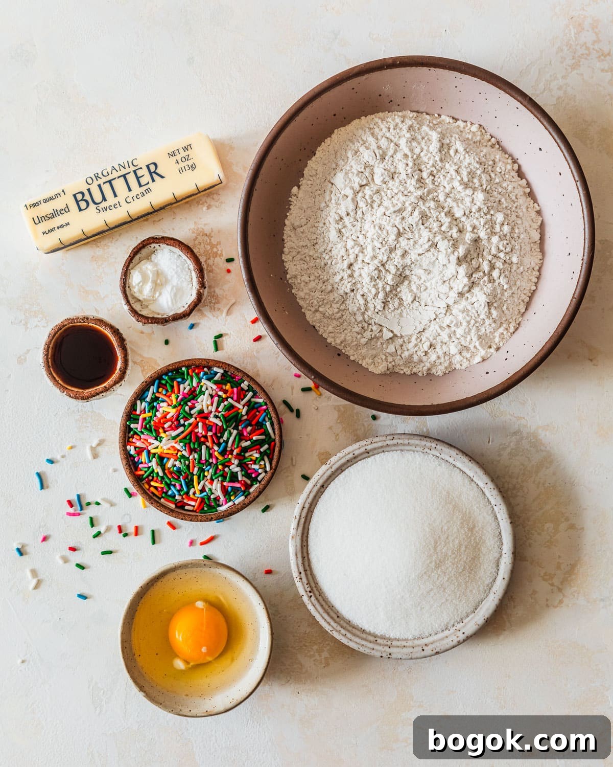Soft and Chewy Mini Sprinkle Cookies Recipe 3 White, brown, and pink bowls of flour, baking soda, salt, vanilla, sugar, an egg, and rainbow sprinkles on a tan counter next to a stick of butter.