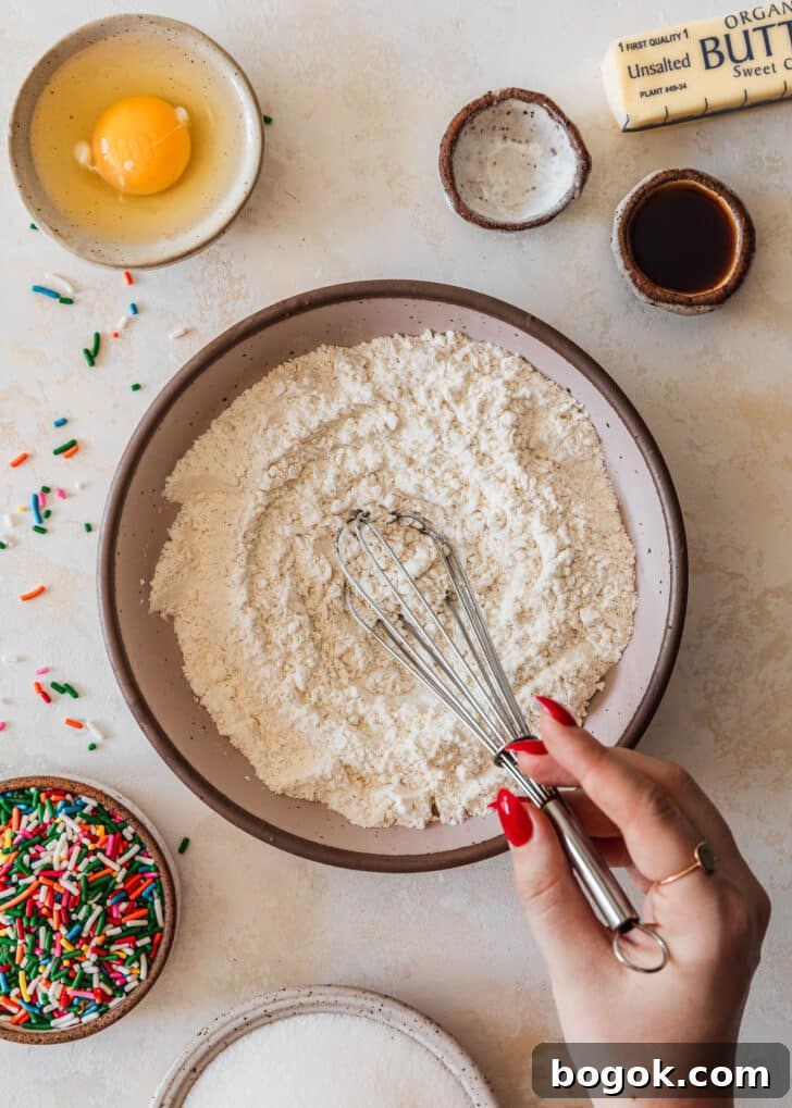 Soft and Chewy Mini Sprinkle Cookies Recipe 4 A hand whisking a pink bowl of flour on a tan counter next to white and brown bowls of sprinkles, sugar, an egg, and vanilla.