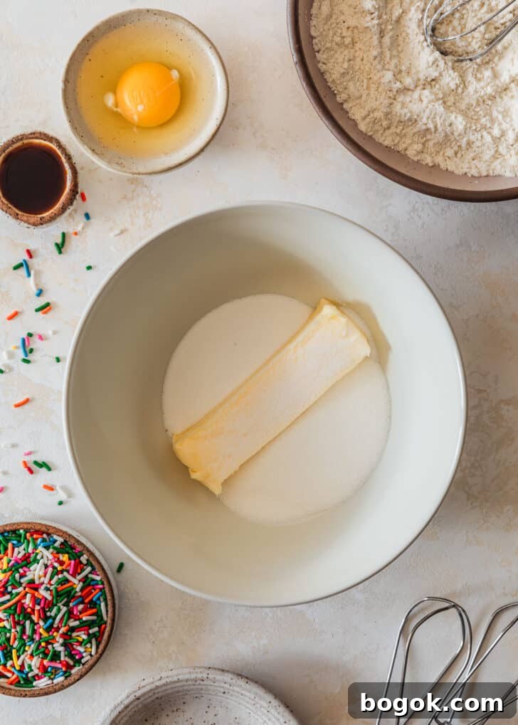 Soft and Chewy Mini Sprinkle Cookies Recipe 5 A white bowl of butter and sugar on a tan counter next to a pink bowl of flour, white bowl with an egg, and brown bowl of sprinkles.