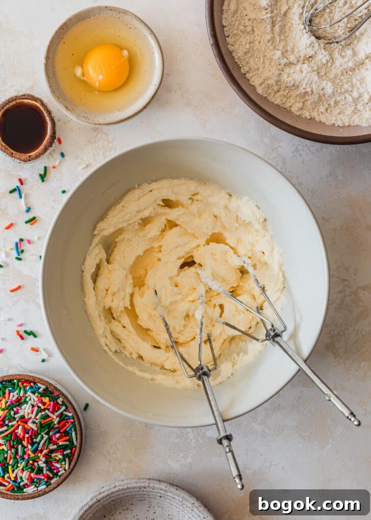 Soft and Chewy Mini Sprinkle Cookies Recipe 6 A white bowl of beaten butter and sugar on a tan counter next to a pink bowl of flour, white bowl with an egg, and brown bowl of sprinkles.