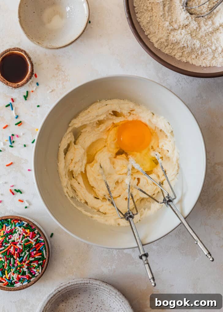 Soft and Chewy Mini Sprinkle Cookies Recipe 7 A white bowl with butter, sugar, and an egg on a tan counter next to a pink bowl of flour, white bowl, and brown bowl of sprinkles.