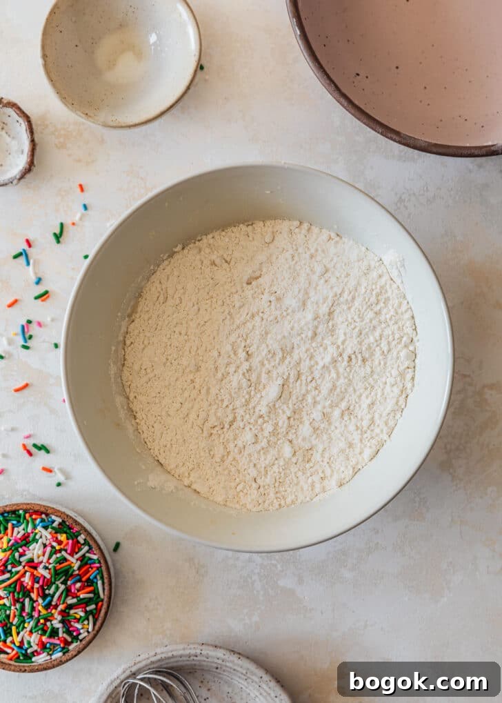 Soft and Chewy Mini Sprinkle Cookies Recipe 8 A white bowl with flour on a tan counter next to a pink bowl, brown bowl of sprinkles, and empty white ingredient bowls.