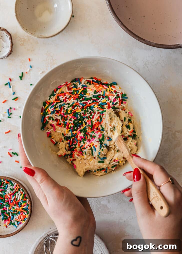 Soft and Chewy Mini Sprinkle Cookies Recipe 9 Hands folding sprinkle cookies dough in a white bowl on a tan counter next to white and pink bowls.