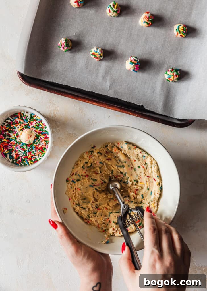 Soft and Chewy Mini Sprinkle Cookies Recipe 10 Hands scooping sprinkle cookies out of a white bowl of dough on a tan counter next to a sheet pan of cookies.